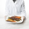 Shef in white jacket holding Classic White Rectangular Serve Platter with steak and carrots on it in front of a white background